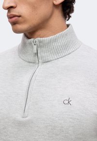 Gros plan d'une personne portant un pull côtelé gris clair à col montant avec une demi-fermeture éclair et un petit logo "ck" sur la poitrine.