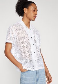 White short-sleeve shirt with a grid pattern and a button-down collar. Fabric appears textured and lightweight, featuring a left chest pocket.
