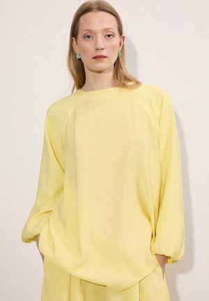 Γυναίκα με ίσια ξανθά μαλλιά, που φοράει πράσινα σκουλαρίκια και άνετο κίτρινο μακρυμάνικο τοπ με ταιριαστή παντελόνα μπροστά σε απλό φόντο.