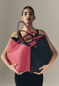 Dos bolsos tote: uno de cuero rosa con textura de grano, otro de cuero negro con textura, ambos con correas alargadas y un pequeño detalle de logotipo.