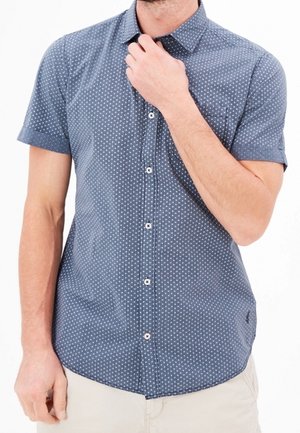 Homme portant une chemise bleue à manches courtes avec un petit motif de points blancs et un pantalon clair, main près du col.