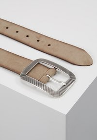 Beiger Wildledergürtel mit einer rechteckigen Schnalle in Silberoptik, die ein strukturiertes Design aufweist und mehrere verstellbare Löcher entlang ihrer Länge hat.