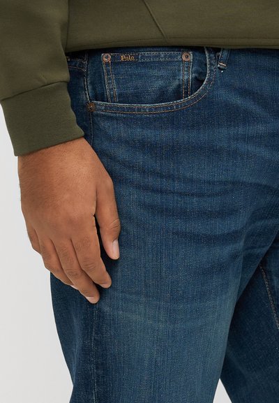 Σκούρο μπλε τζιν παντελόνι με διακριτική υφή, εμφανείς ραφές και μπροστινή τσέπη. Μερικώς ορατό μανίκι σε ελιά πράσινο χρώμα.