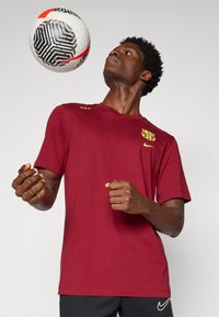 Kohvamaroon Nike särk lühikeste varrukatega, millel on kollane logo ja aktsendid, seljas inimesel, kes jongleerib must-valge mustriga jalgpalliga.