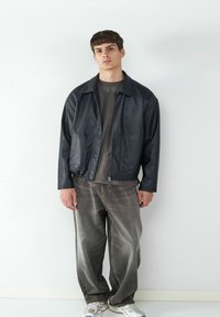 Giacca nera in pelle con zip e due tasche anteriori, abbinata a jeans grigi larghi e sneakers chiare. Sfondo uniforme, senza pattern visibili.