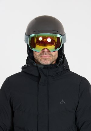 Man draagt een zwarte skihelm, reflecterende skibril en een zwarte winterjas, kijkt recht vooruit tegen een effen achtergrond.