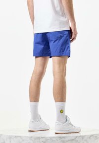 Blå svømmeshorts med let tekstur, sidelomme og elastisk talje. Sammen med hvide sneakers og sokker med smiley ansigt.