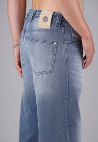Man wearing light blue jeans with back pockets and a tan brand label on the waistband, standing against a plain gray background.