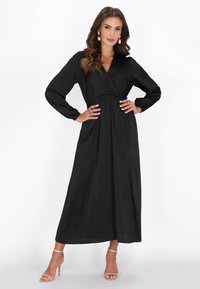 Schwarzes Langarmkleid mit Wickeldesign, tailliert und mit fließendem Rock. Kombiniert mit beigen Riemchenabsätzen und hängenden Ohrringen.