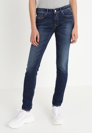 Slim-fit dark blue jeans worn with a gray top and white sneakers on a standing person against a plain white background.