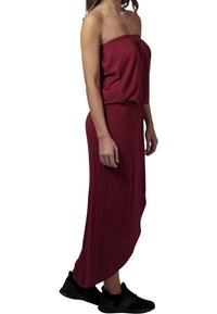 Viininpunainen olkaimeton maximekko, jossa on kerätty kaula-aukko, virtaava muotoilu ja pehmeä tekstuuri, yhdistettynä mustiin urheilukenkiin.