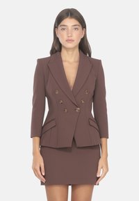 Blazer marrone doppiopetto con bottoni dorati, vestibilità su misura, revers sottili, maniche tre quarti e tasche frontali. Abbinato a una gonna coordinata.