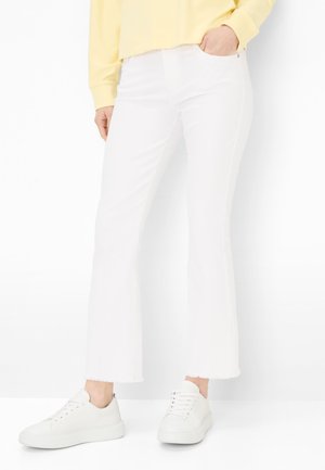 Persona che indossa jeans bianchi a zampa leggermente corti, sneakers bianche e una maglia gialla a maniche lunghe, con una mano in tasca.