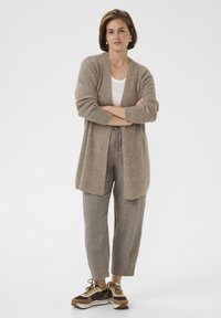 Femme debout les bras croisés, portant un cardigan beige, un pantalon marron clair, une chemise blanche et des baskets marron sur fond blanc.
