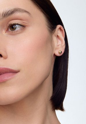 Złoty kolczyk w kształcie sztyftu na uchu modelki, o zaokrąglonym kształcie i gładkiej fakturze. Tło jest neutralne, aby podkreślić kolczyk.