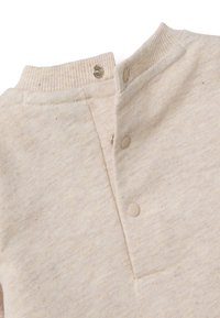 Licht beige katoenen pullover met een geribde kraag, drie drukknoopjes en een klein zakje. Zachte textuur met subtiele kleurvariaties.