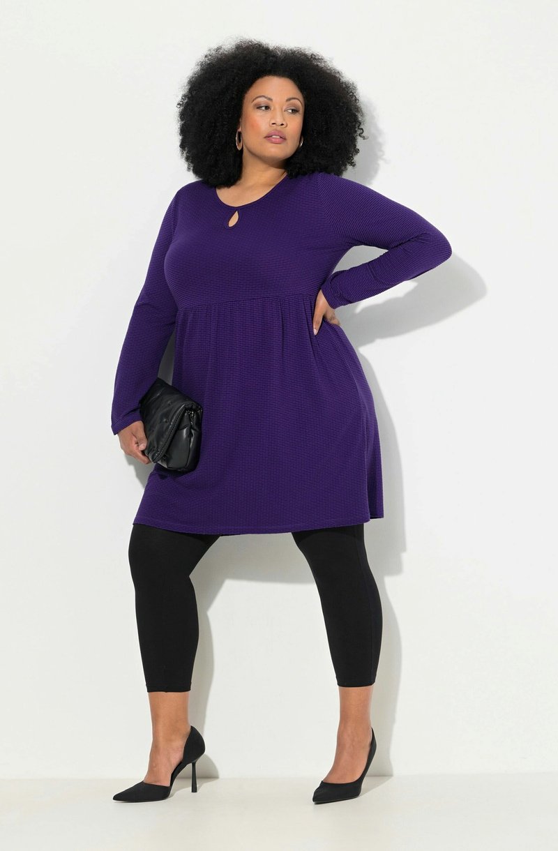 Frau mit lockigem Haar, die ein lila-langärmliges Kleid, schwarze Leggings, schwarze High Heels trägt und eine schwarze Clutch-Tasche hält.