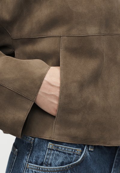 Χέρι μερικώς μέσα στην πλαϊνή τσέπη ενός καφέ σουέτ μπουφάν που φοριέται πάνω από μπλε τζιν παντελόνια.