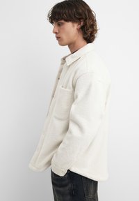 Valkoinen, kuvioitu fleece-paitatakki, jossa kaulus, etutasku ja napinlävet. Yllä tummien farkkujen kanssa, sivuprofiilissa.