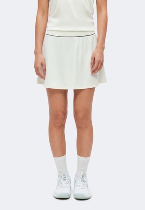 Vrouw draagt een korte witte plissérok, witte top, witte sokken en witte sportschoenen, staand met armen ontspannen langs het lichaam.