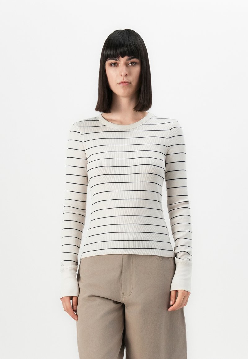 Jonge vrouw met steil donker haar, gekleed in een witte lange mouwtop met dunne zwarte strepen en beige broek, staande tegen een effen achtergrond.