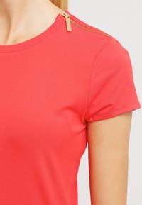 Korallfärgad t-shirt med kort ärm gjord av ett mjukt tyg, med en guldig dragkedjedetalj på axeln och en klassisk rundad halsringning.