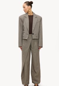 Mujer con blazer oversize marrón de cuadros y pantalones de pierna ancha, lleva pendientes de aro dorados y una camiseta marrón oscuro, de pie frente a un fondo blanco.