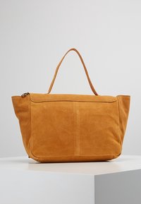 Sac fourre-tout en daim jaune moutarde avec une anse unique, une fermeture éclair et une texture douce. Présente une surface lisse et un design minimaliste.