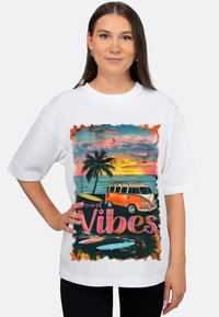 Witte oversized t-shirt met een kleurrijke afbeelding van een vintage busje, palmbomen en zonsondergang, met de tekst "Good Vibes" in roze.