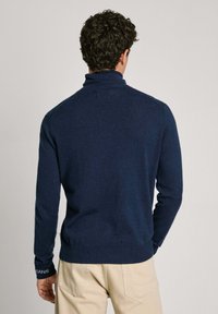 Tumši zila adīta turtlenecks džemperis ar garām piedurknēm, ar pieguļošu dizainu un ribinātām aprocēm, uzvilkts virs gaišas krāsas biksēm.