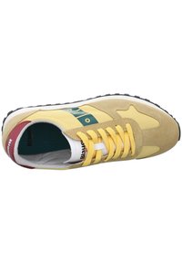 Sneaker giallo e beige con lacci gialli, brandizzazione verde e bianca sul lato e un dettaglio rosso sul tallone, mostrato visto dall'alto.