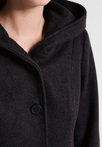 Primo piano di una persona che indossa un cappotto di lana grigio scuro con un ampio cappuccio e due bottoni neri visibili chiusi sul davanti.