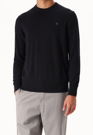 Homme portant un pull noir ajusté à manches longues et col rond avec un petit logo sur la poitrine gauche et un pantalon gris clair, mains partiellement dans les poches.