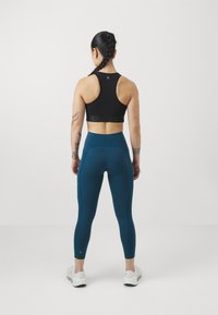 Reggiseno sportivo nero con una fascia elastica marchiata, abbinato a leggings teal a vita alta con tasche e cuciture a contrasto. Scarpe bianche.