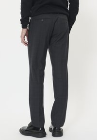 Sötétszürke kockás nadrágot viselő férfi, hátul gombos zsebekkel, fekete fényes Dress cipőkkel, fehér háttér előtt áll.