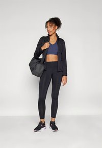 Czarna kurtka zapinana na zamek, granatowy biustonosz sportowy, czarne legginsy oraz czarne sneakersy z fioletowymi akcentami. Nosi szarą torbę sportową. Gładka tekstura tkaniny.