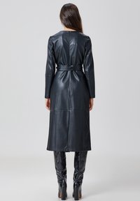 Manteau long en cuir noir avec une taille ceinturée, des manches longues et une finition lisse. Comprend une fermeture éclair au dos et un design avec des coutures en panneaux.