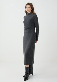 Graues Strickkleid mit langen Ärmeln und steigendem Kragen, mit einer seitlichen Knotendetail und geradem Schnitt. Trägt dazu spitze schwarze Stiefeletten bis zum Knöchel.