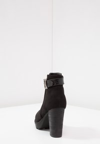 Botte de cheville en suede noir avec une sangle texturée noire et une boucle en argent, dotée d'un talon épais en bois et d'un design à bout rond.
