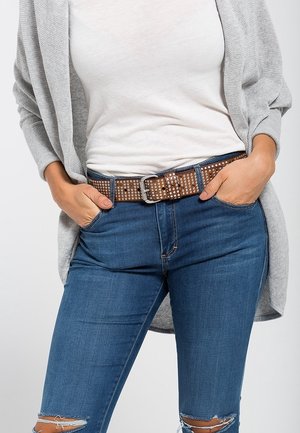 Femme portant un haut blanc, un cardigan gris, un jean bleu déchiré et une ceinture marron cloutée, les mains dans les poches.