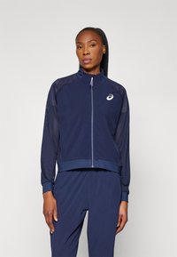 Marineblå sportsjakke med lynlås, meshærmer og krave, med et hvidt logo på brystet og ribbekantede manchetter. Struktureret design hele vejen igennem.