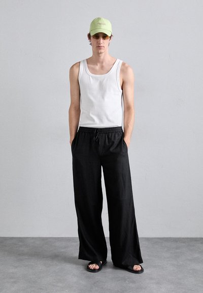 Top sin mangas blanco, pantalones negros holgados y sandalias negras; lleva una gorra verde claro y gafas de sol con tinte amarillo; diseño minimalista, estilo casual.