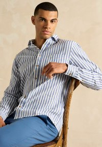 Zila un balta svītrota pogcauruma krekls, kas izgatavots no auduma, ar krūšu kabatu un koka pogām, nēsāts ar zilām biksēm.