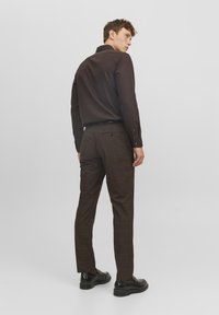 Camicia marrone a bottoni con una vestibilità slim, abbinata a pantaloni a quadri marroni e scarpe nere, mostrata da dietro su uno sfondo semplice.
