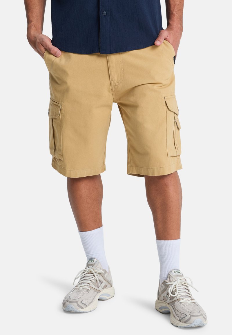 Pantaloni cargo beige con più tasche laterali, taglio dritto, realizzati in materiale misto cotone, abbinati a calzini bianchi e sneakers chiare.