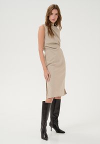 Robe beige sans manches avec une silhouette ajustée et des fentes sur le côté, associée à des bottes noires à talons hauts en cuir brillant.