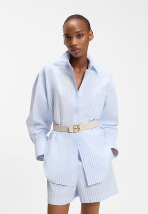 Kvinde iført lyseblå og hvide stribede, oversize skjorte og matchende shorts, accessoriseret med et bredt beige bælte, stående med hænderne i lommerne.