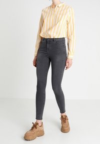 Jeans skinny a vita alta grigi abbinati a una camicia a righe gialle e bianche con bottoni e sneakers chunky marroni con lacci.