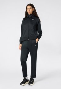 Czarny sportowy dres składający się z kurtki zapinanej na zamek z wysokim kołnierzem oraz dopasowanych spodni, obydwa z akcentami logo. Całość dopełniają sneakersy.