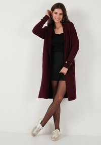 Tânără zâmbitoare, îmbrăcată cu un cardigan lung de culoare burgundă, un top negru, pantaloni scurți negri, ciorapi transparenți și teniși albi, stând în fața unei pereți albi.